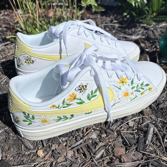 A pair of white low-top sneakers with yellow heel tabs, decorated with hand-painted yellow daisies, green leaves, and bumblebees, resting on a bed of mulch outdoors.