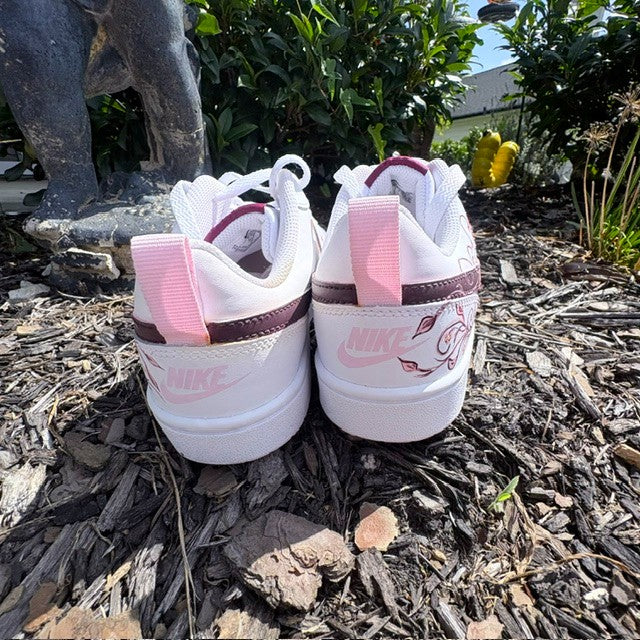 A pair of white Nike low-top sneakers shown from the back, placed on a mulch surface outdoors with greenery and a statue in the background. The shoes feature pink pull tabs, pink Nike branding, and a dark burgundy stripe near the collar.