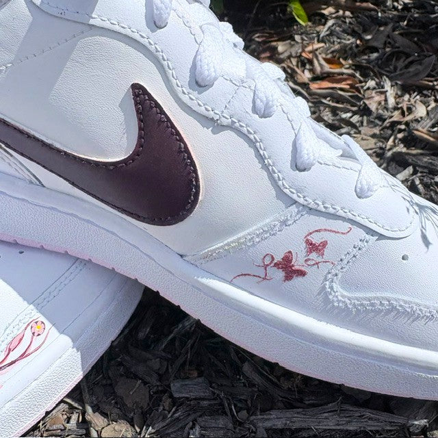 White Nike low-top sneaker with a dark brown Swoosh and red embroidered script on the side, resting on a bed of dark mulch outdoors.