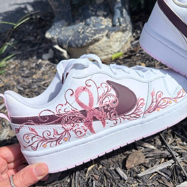 A white low-top sneaker with a pink awareness ribbon and floral scroll design on the side, held by a hand outdoors on mulch. The shoe has a dark mauve Nike Swoosh and pink laces.