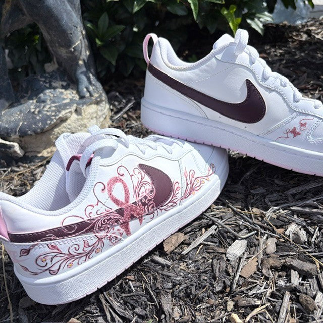 A pair of white Nike low-top sneakers with dark burgundy swooshes and pink ribbon graphics, displayed on a mulch surface outdoors.