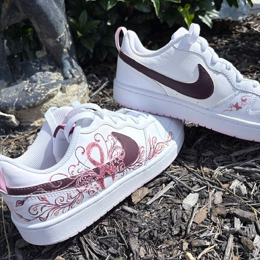 A pair of white Nike low-top sneakers with dark burgundy swooshes and pink ribbon graphics, displayed on a mulch surface outdoors.