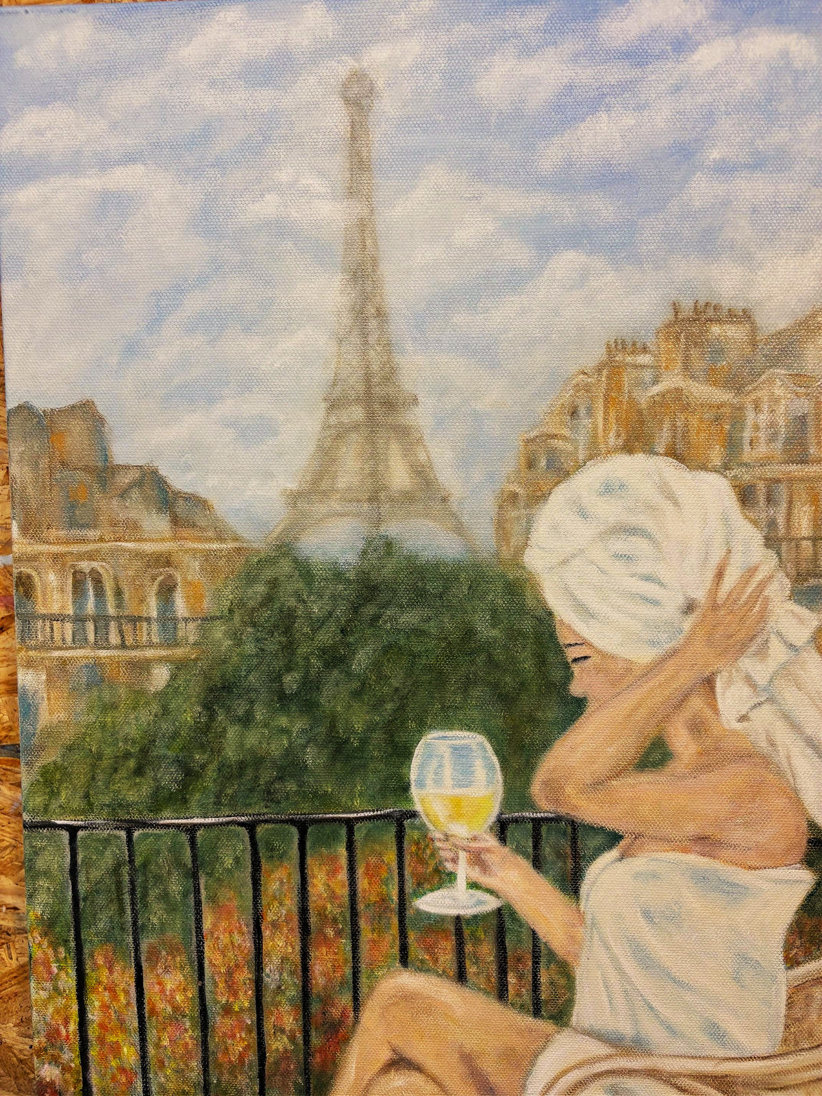 A painted scene of a woman wrapped in white towels sitting on a balcony and holding a glass of white wine, with the Eiffel Tower and Parisian buildings in the background.